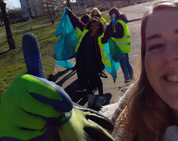 Scholier met werkhandschoen steekt duim omhoog in selfie met op de achtergrond klasgenoten die zwerfafval opruimen